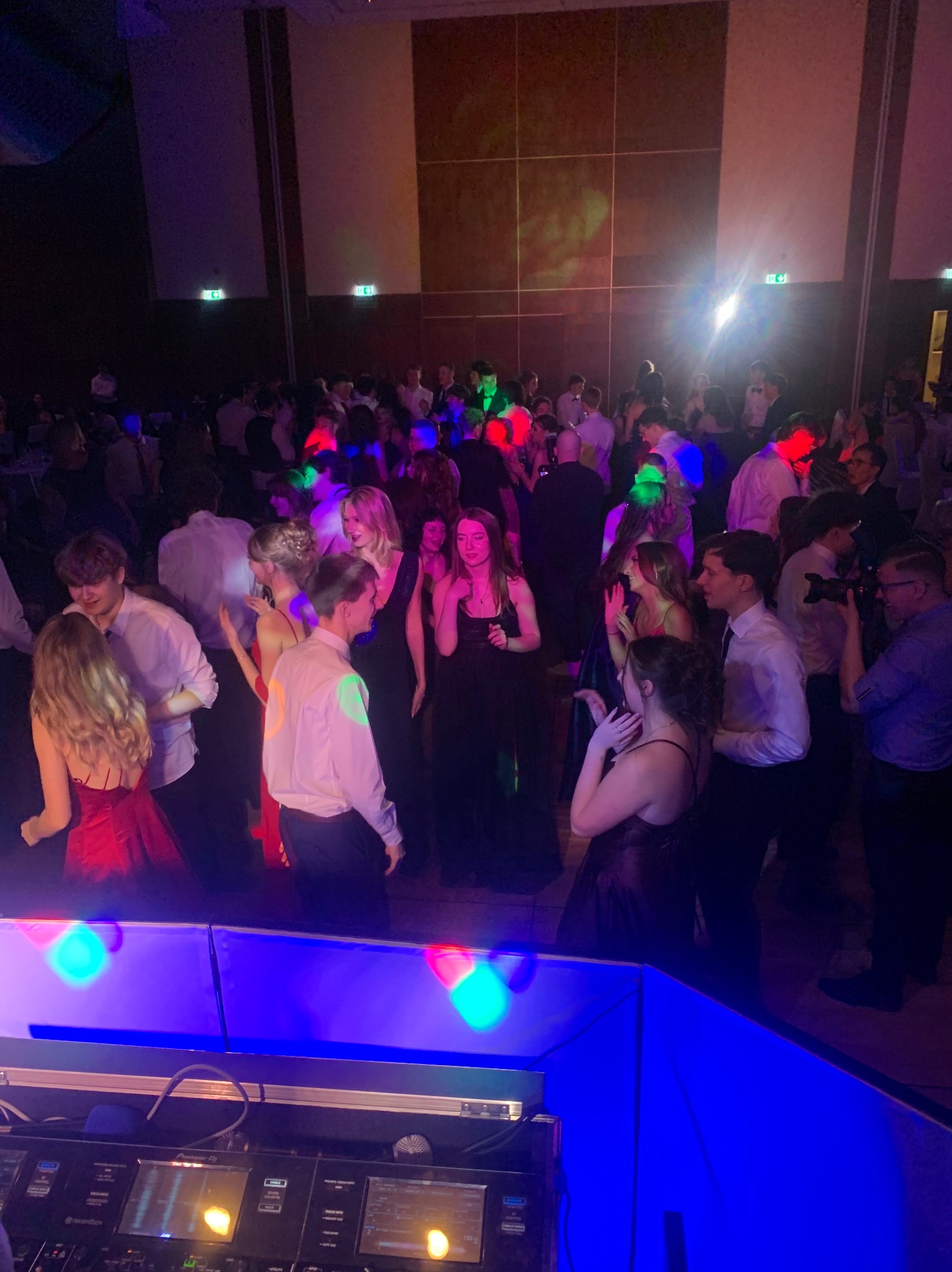 Parkiet taneczny wypełniony tańczącymi ludźmi, oświetlony kolorowymi światłami dyskotekowymi, z widocznym stanowiskiem DJ-a w dolnej części kadru.