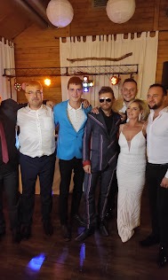 Zespół muzyczny w kompletnym składzie pozuje do pamiątkowego zdjęcia z parą młodą i gośćmi weselnymi na tle drewnianej sali z lampionami.