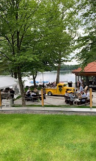 Plenerowa impreza nad jeziorem z żółtym, stylizowanym samochodem jako atrakcją. Goście siedzą przy stolikach, w tle woda i drzewa.