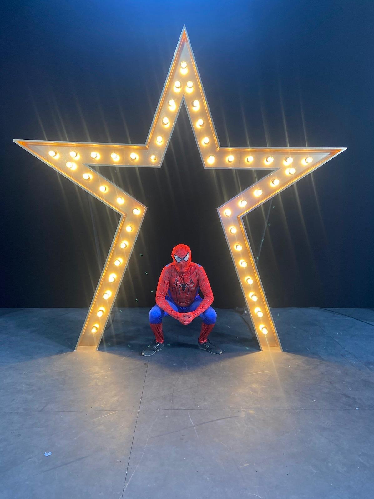 Mężczyzna w kostiumie Spider-Mana kuca przed dużą, podświetlaną gwiazdą na czarnym tle. Ujęcie z dołu.