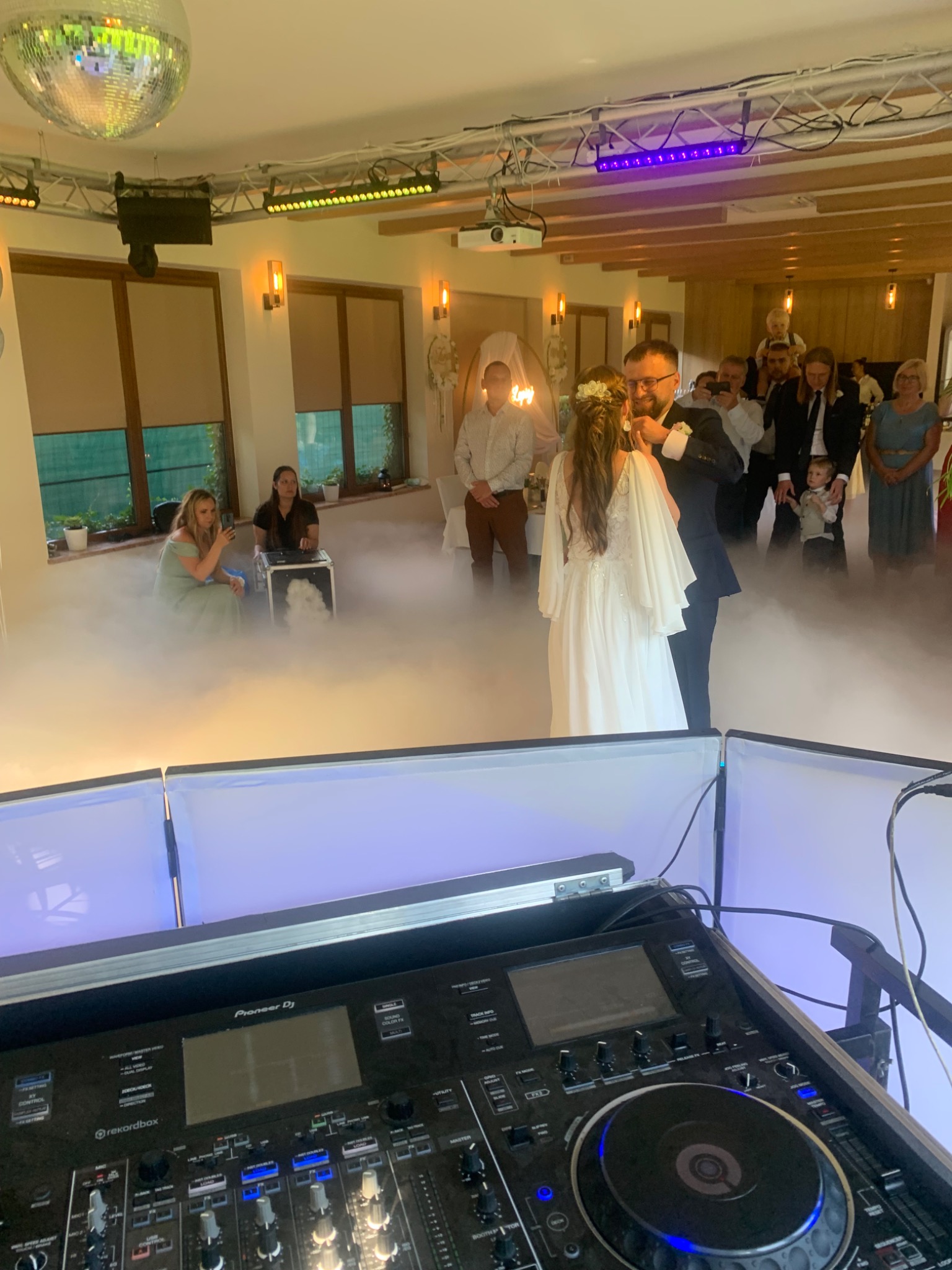 Para młoda tańczy w oparach dymu na sali weselnej z oświetleniem i sprzętem DJ-skim na pierwszym planie. Dekoracje i goście w tle.
