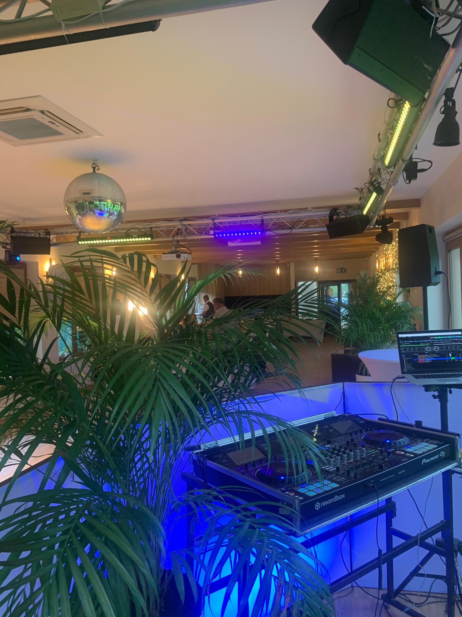 Sala z DJ-ką Pioneer DJ, oświetleniem estradowym i kulą dyskotekową. Dekoracje z roślin doniczkowych. Scena gotowa na imprezę.