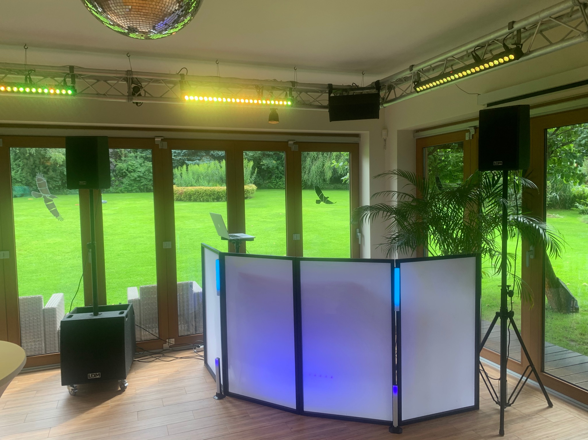 Stanowisko DJ'a z białym podświetleniem, nagłośnieniem i oświetleniem na tle okien z widokiem na zielony ogród. Na suficie kula dyskotekowa.