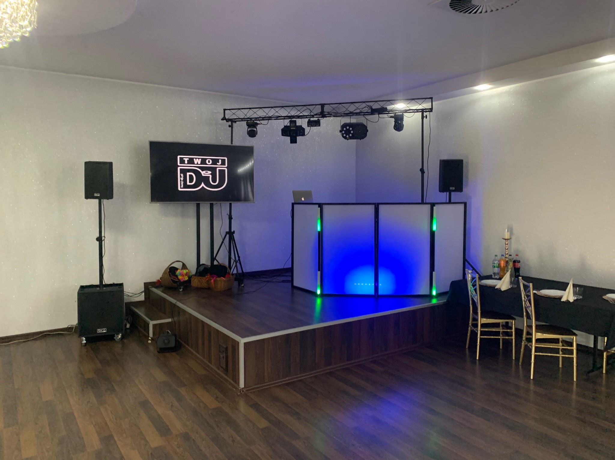 Scena DJ-a z oświetleniem i nagłośnieniem gotowa na imprezę. Biały panel frontowy podświetlony na niebiesko, głośniki po bokach, ekran z logo DJ-a.
