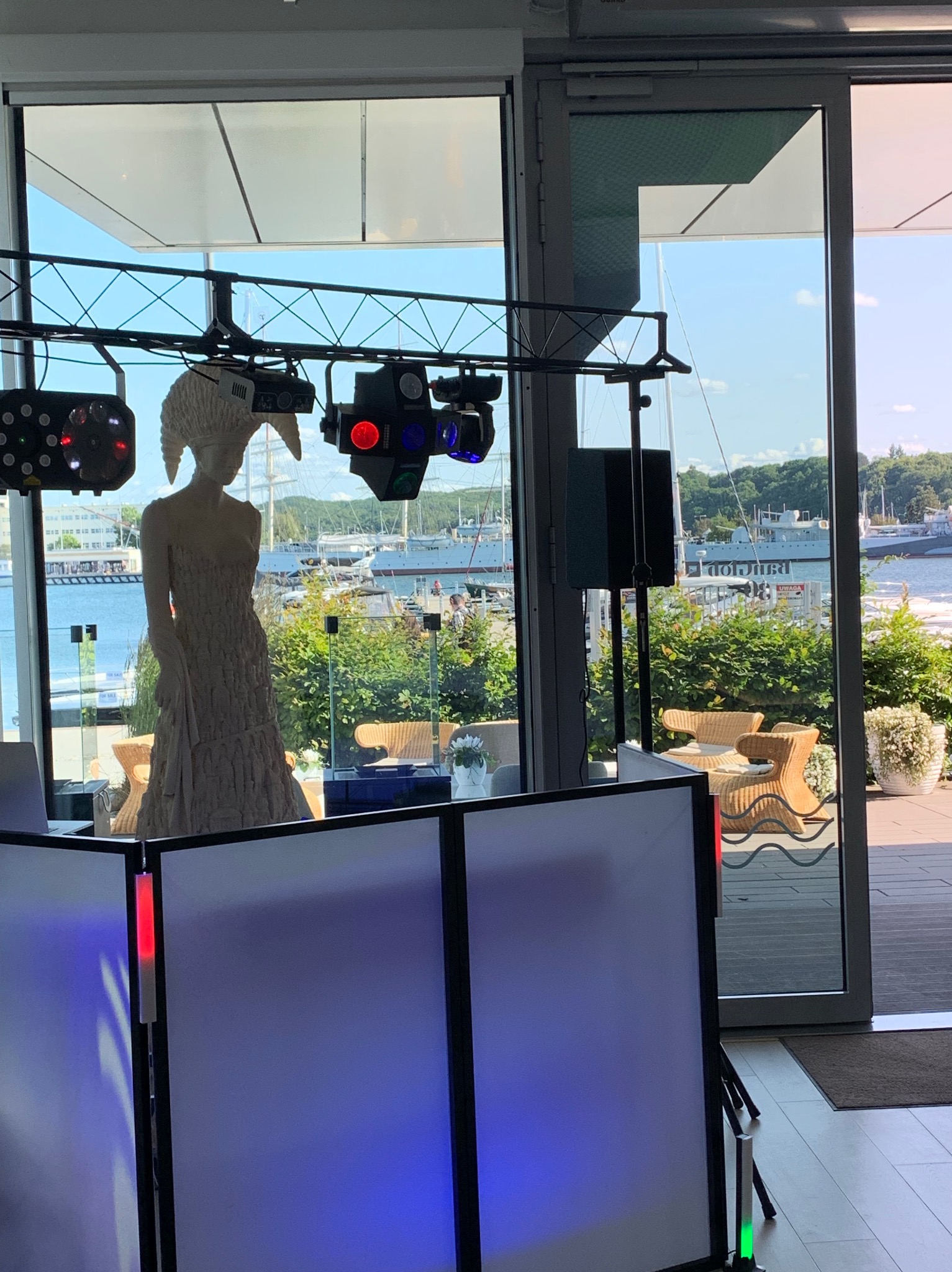 Dekoracja rzeźbiarska pod oświetleniem eventowym, widok na port i morze przez przeszkloną ścianę. Scena z konsoletą DJ-ską w klubie.