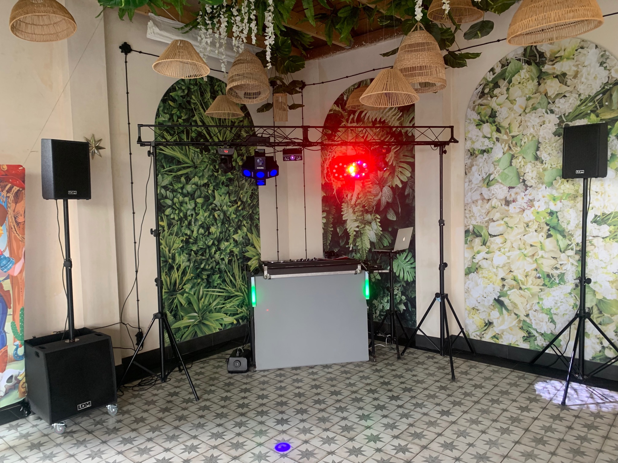 Rozstawiony sprzęt DJ-ski z oświetleniem na tle dekoracyjnych ścian z motywami roślinnymi i kwiatowymi. Widoczne głośniki, konsoleta i lampy w stylu boho.