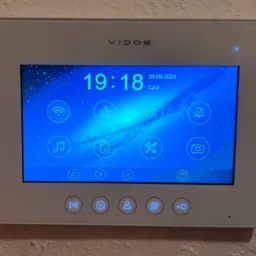 Biały panel sterowania wideodomofonu Vidos z wyświetlaczem pokazującym godzinę 19:18, datę 28-09-2023 i ikonami funkcji, zamontowany na ścianie o szarej fakturze.