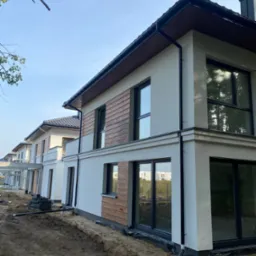 Nowoczesne domy w zabudowie szeregowej z elewacją w jasnym tynku i elementami drewnopodobnymi, widoczne rury spustowe i przygotowany teren wokół budynków.