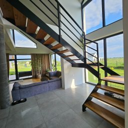 GoHouse Usługi spawalniczo ślusarskie - Nowoczesne schody z czarną metalową konstrukcją i drewnianymi stopniami w przestronnym wnętrzu domu z dużymi oknami wychodzącymi na zielony ogród.