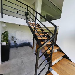 GoHouse Usługi spawalniczo ślusarskie - Nowoczesne schody z drewnianymi stopniami i czarną metalową konstrukcją, widok z dołu, w tle ściana z dekoracyjnym malowaniem przypominającym krajobraz.
