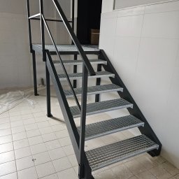 GoHouse Usługi spawalniczo ślusarskie - Metalowe schody z kratowymi stopniami, pomalowane na czarno, w pomieszczeniu z białymi kafelkami na ścianach i podłodze. Widoczne ślady prac wykończeniowych.