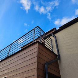 GoHouse Usługi spawalniczo ślusarskie - Nowoczesna balustrada balkonowa z prostych, czarnych profili stalowych, zamontowana na budynku z brązową elewacją imitującą drewno, widok z dołu na tle błękitnego nieba z chmurami.