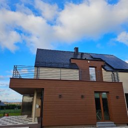 GoHouse Usługi spawalniczo ślusarskie - Nowoczesny dom z brązową elewacją, balkonem z prostą balustradą nierdzewną i czarnym dachem, widok z dołu na tle błękitnego nieba z chmurami.