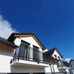 Nowoczesna balustrada balkonowa z czarnych, pionowych prętów na tle białej elewacji budynku i błękitnego nieba z chmurami.