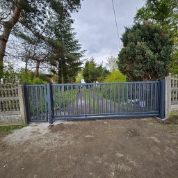 GoHouse Usługi spawalniczo ślusarskie - Nowoczesna, przesuwna brama wjazdowa z pionowych, szarych profili, zamontowana między betonowymi słupkami z dekoracyjnym ażurem, na tle zieleni drzew i pochmurnego nieba.
