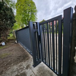 GoHouse Usługi spawalniczo ślusarskie - Nowoczesna, częściowo otwarta, przesuwna brama wjazdowa w kolorze grafitowym, z pionowymi szczeblami, zamontowana na betonowym podłożu, z widocznym mechanizmem otwierania i zielenią w tle.