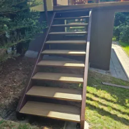 Zewnętrzne schody z brązową metalową ramą i deskami kompozytowymi, prowadzące na balkon domu otoczonego zielenią.