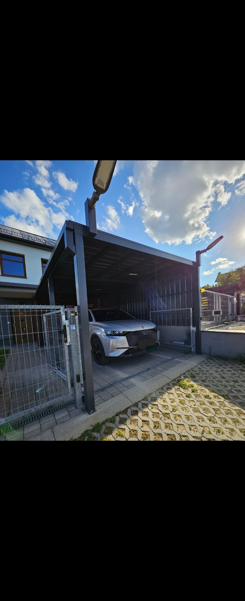 Nowoczesna wiata garażowa z metalową konstrukcją w kolorze antracytowym, częściowo osłonięta metalowymi panelami, z zaparkowanym srebrnym samochodem osobowym i przesuwną bramą wjazdową z metalowej...