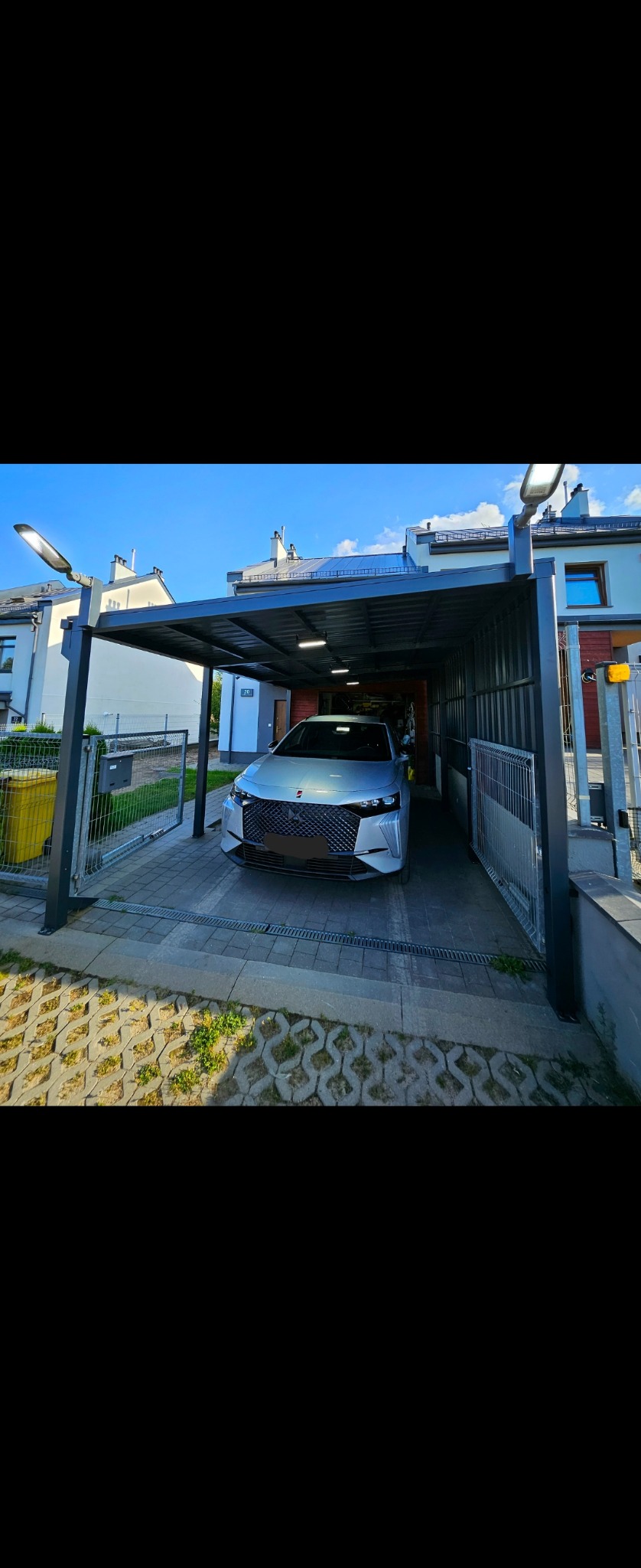 Nowoczesny carport z szarej stali z oświetleniem LED, chroniący srebrny samochód na betonowej kostce brukowej.