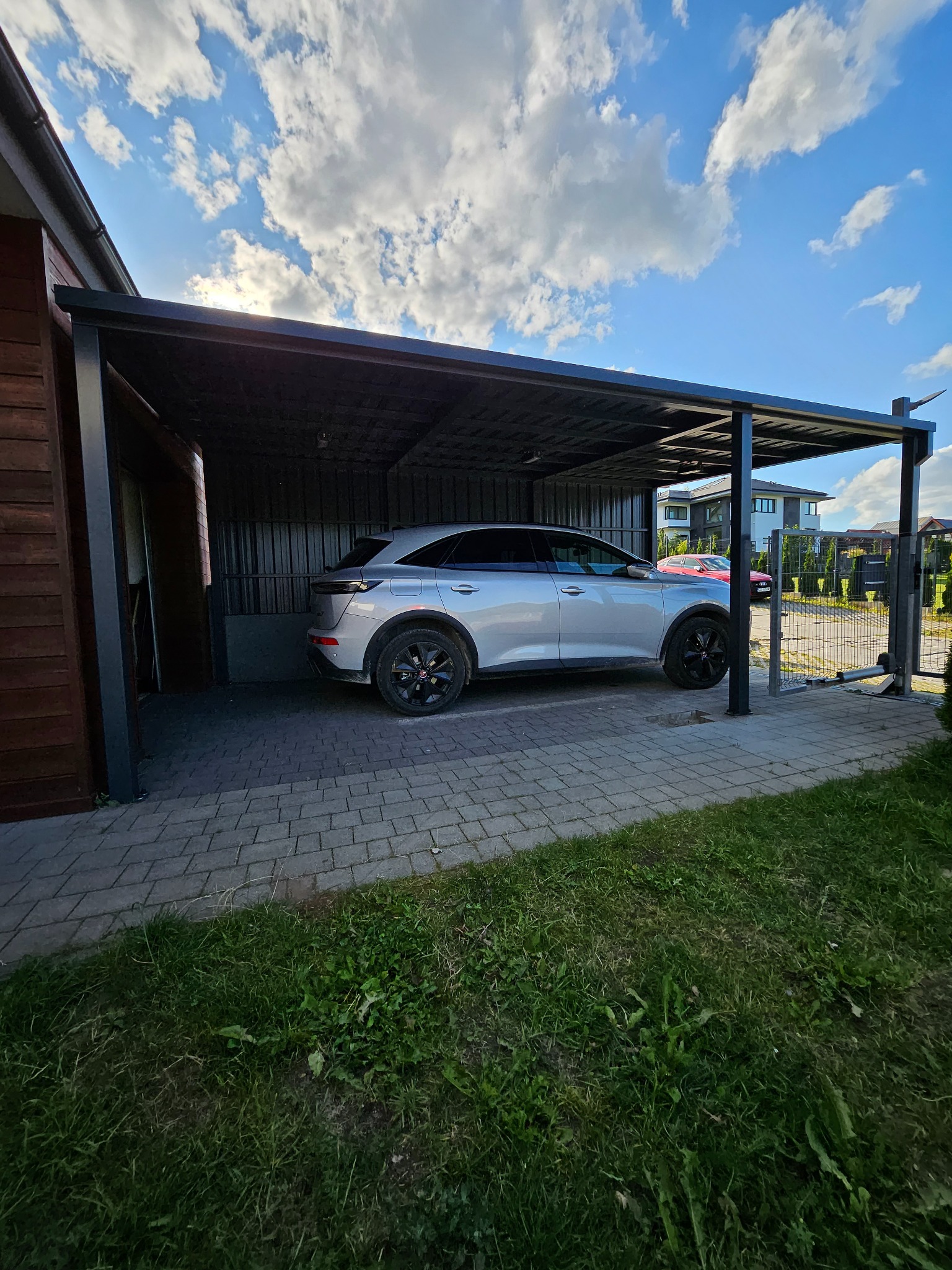 Zadaszenie metalowe dla samochodu w Łapalicach, przylegające do drewnianego budynku, z widocznym srebrnym SUV-em i fragmentem ogrodzenia panelowego w tle.