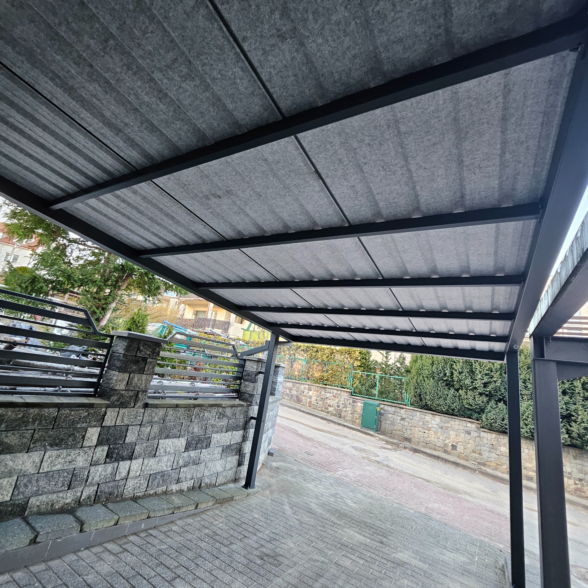 Nowoczesna wiata garażowa z szarą, falistą blachą dachową i czarną, metalową konstrukcją, przylegająca do kamiennego murku z metalowym ogrodzeniem.