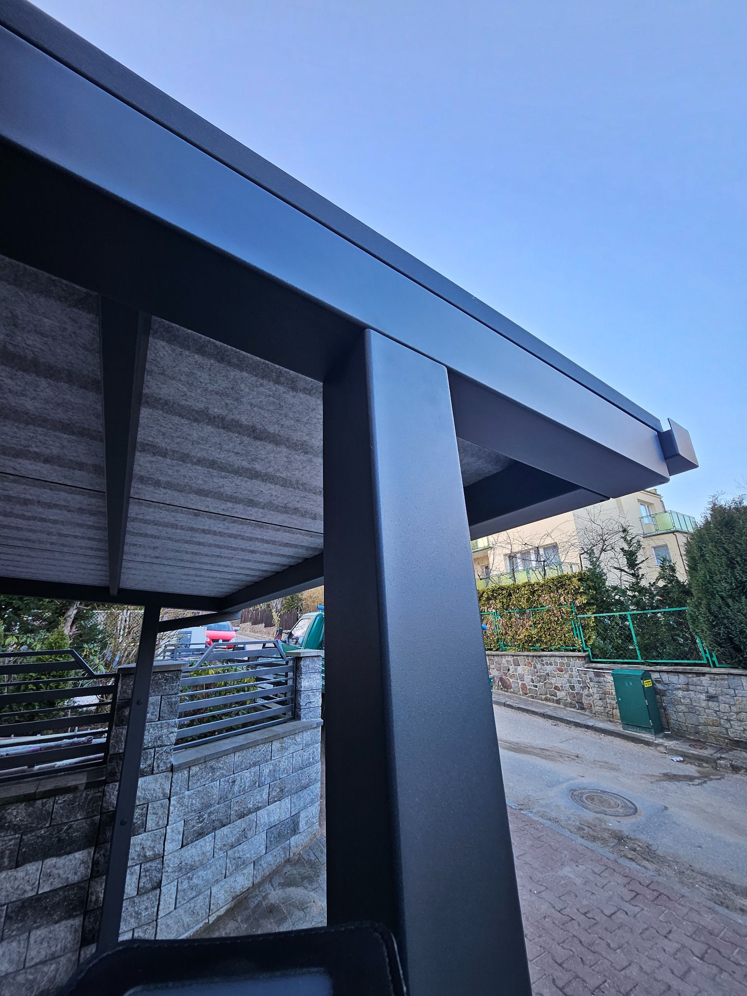 Nowoczesna, czarna konstrukcja zadaszenia nad podjazdem, widok od dołu, z szarym podsufitką i elementami ogrodzenia w tle.