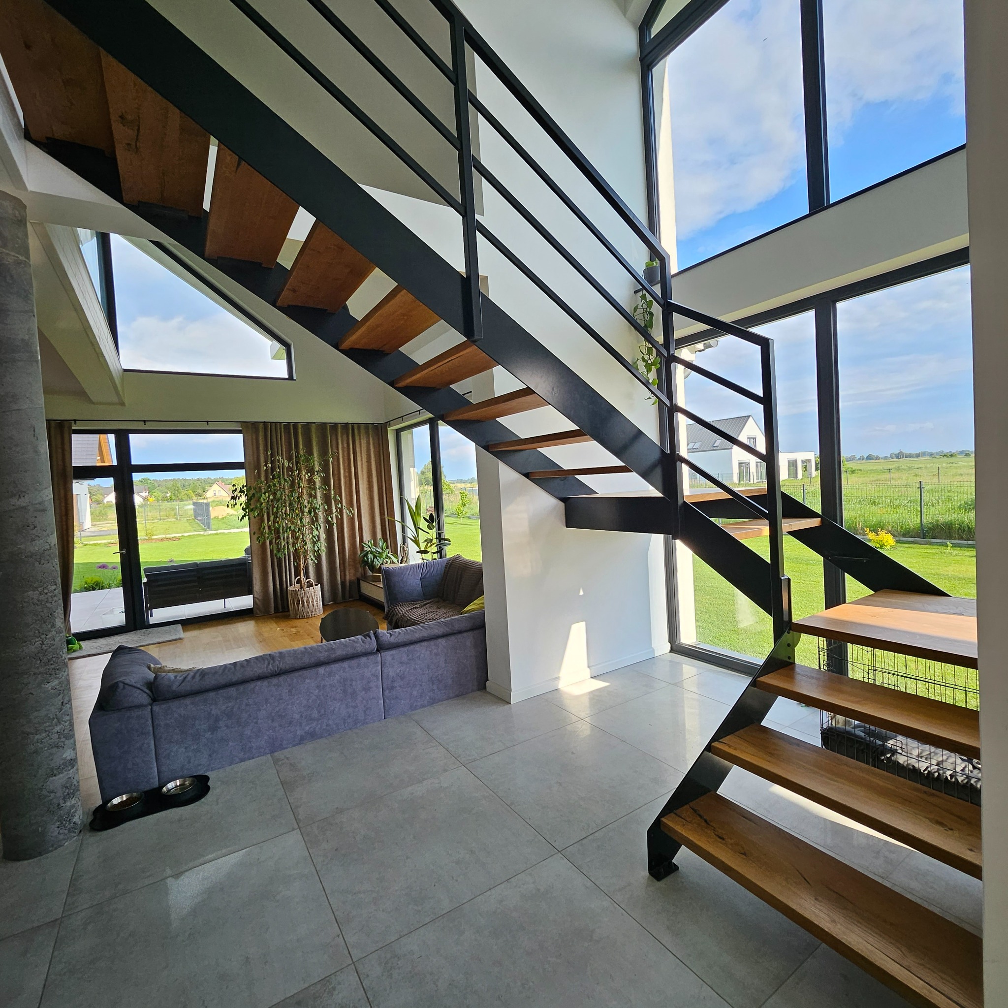 Nowoczesne schody z czarną metalową konstrukcją i drewnianymi stopniami w przestronnym wnętrzu domu z dużymi oknami wychodzącymi na zielony ogród.