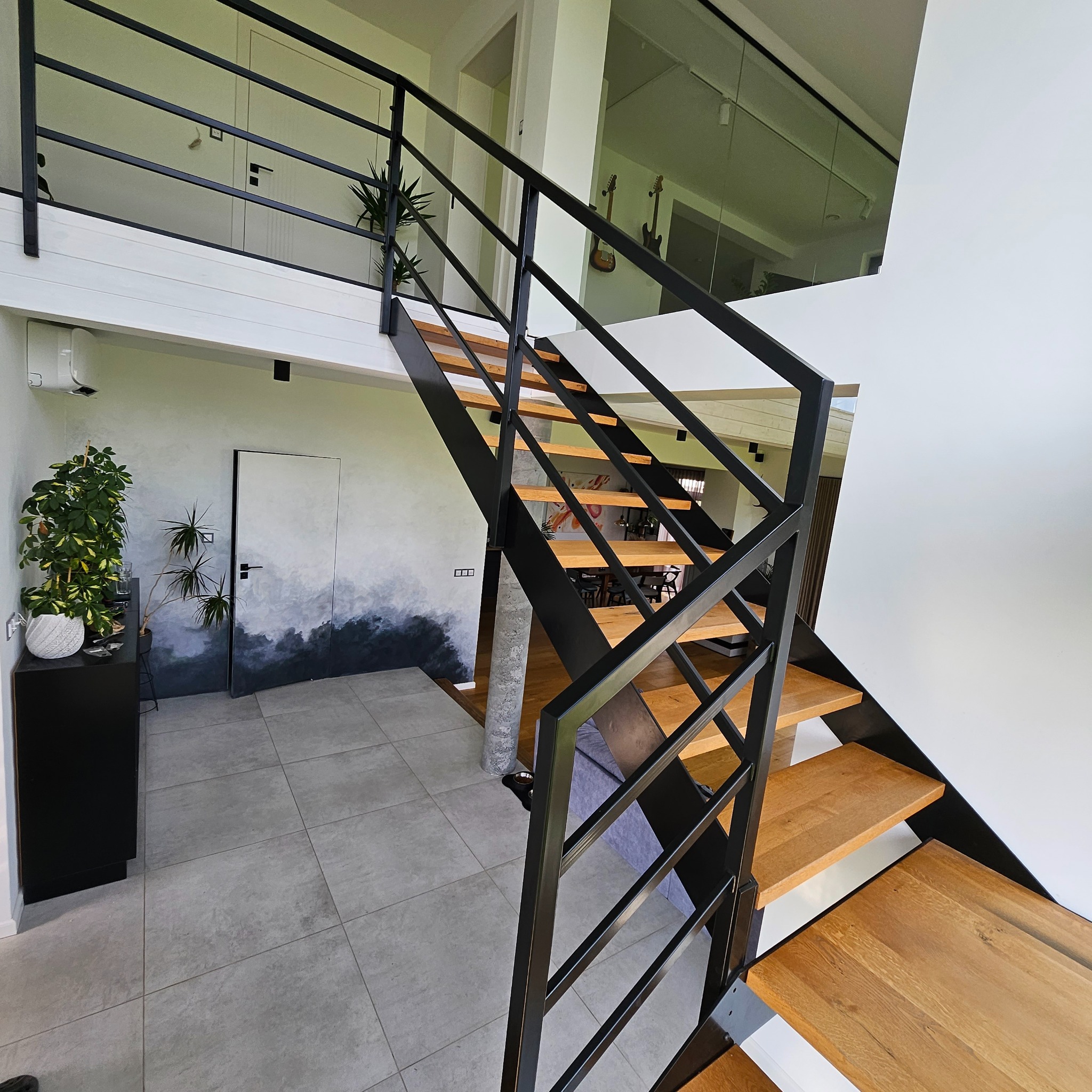 Nowoczesne schody z drewnianymi stopniami i czarną metalową konstrukcją, widok z dołu, w tle ściana z dekoracyjnym malowaniem przypominającym krajobraz.