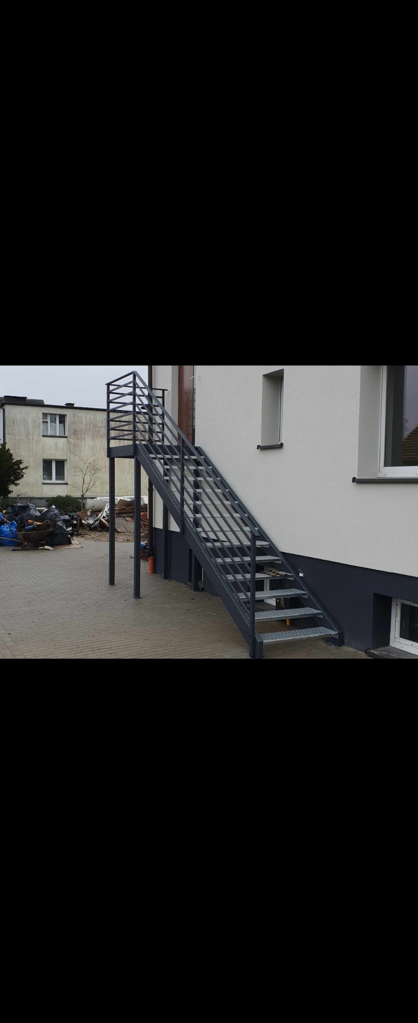 Zewnętrzne schody metalowe z ażurową balustradą prowadzące do drzwi na piętrze budynku, widoczne metalowe stopnie z kratki.