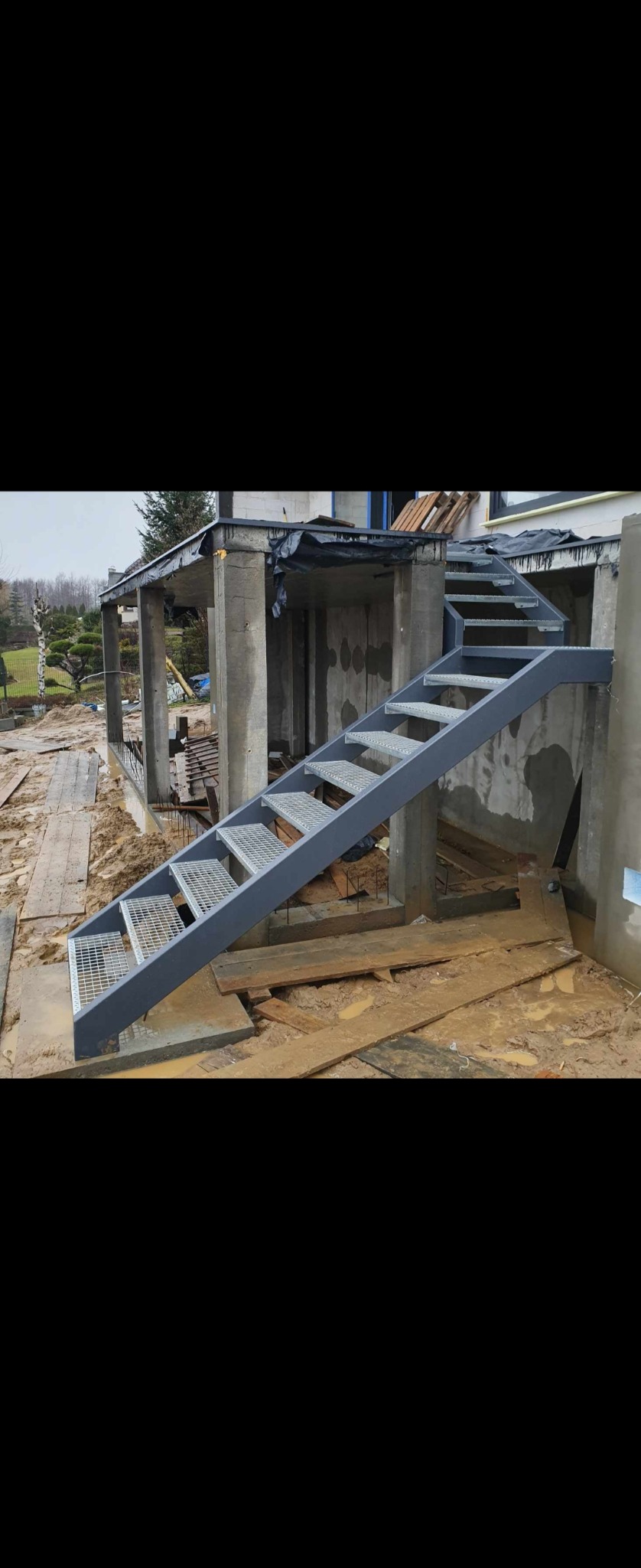Zewnętrzne schody z metalową kratownicą prowadzące do surowego budynku w trakcie budowy, z widocznym błotem i elementami konstrukcyjnymi.