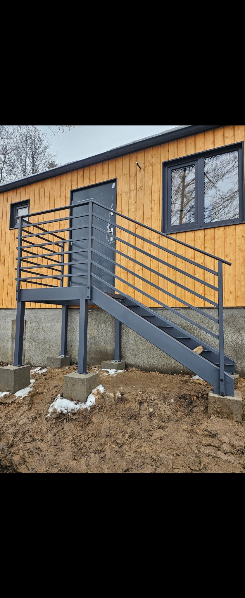 Zewnętrzne schody z metalową balustradą w kolorze grafitowym, prowadzące do drzwi wejściowych drewnianego domu, osadzone na betonowych fundamentach w surowym terenie.