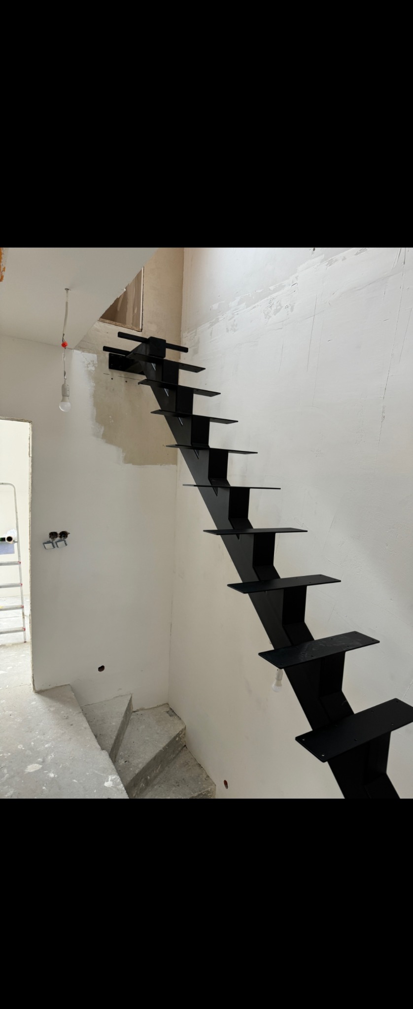 Nowoczesne, czarne, metalowe schody wspornikowe zamontowane w surowym wnętrzu w trakcie remontu. Widoczne niedokończone ściany i betonowe stopnie na dole.