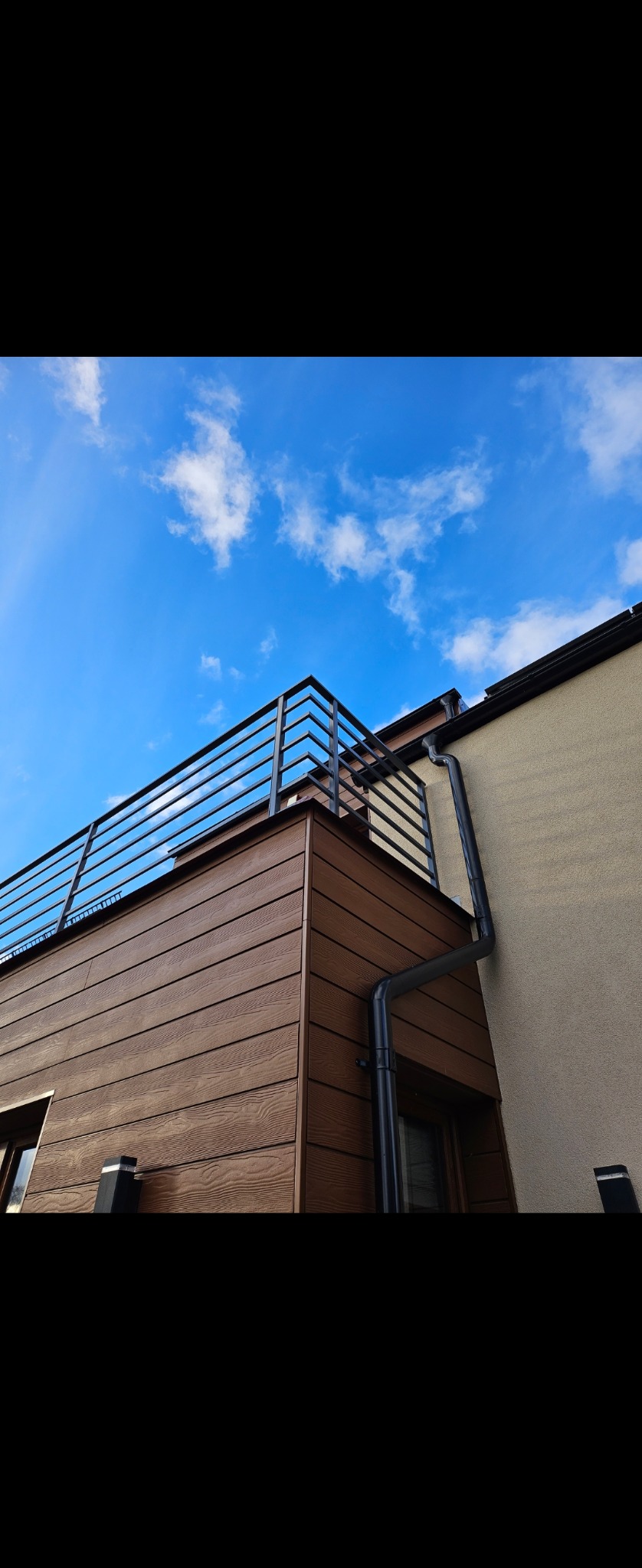 Nowoczesna balustrada balkonowa z prostych, czarnych profili stalowych, zamontowana na budynku z brązową elewacją imitującą drewno, widok z dołu na tle błękitnego nieba z chmurami.