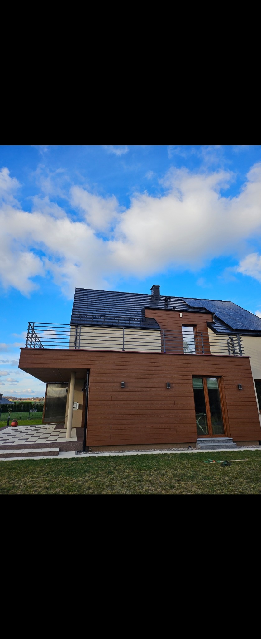 Nowoczesny dom z brązową elewacją, balkonem z prostą balustradą nierdzewną i czarnym dachem, widok z dołu na tle błękitnego nieba z chmurami.
