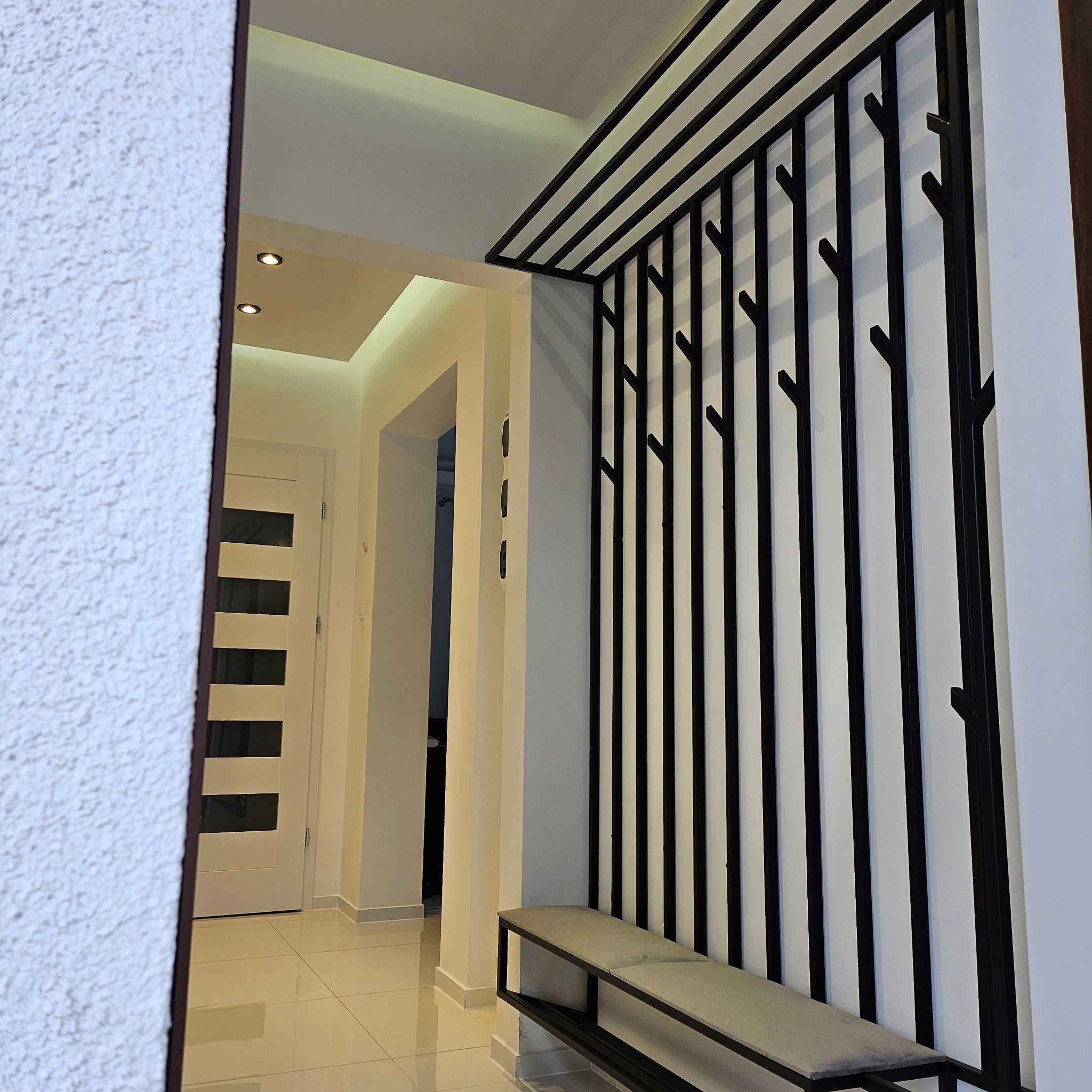 Nowoczesny przedpokój z metalową konstrukcją w formie wieszaka z siedziskiem, czarne pionowe elementy na białym tle, widok z zewnątrz pomieszczenia.