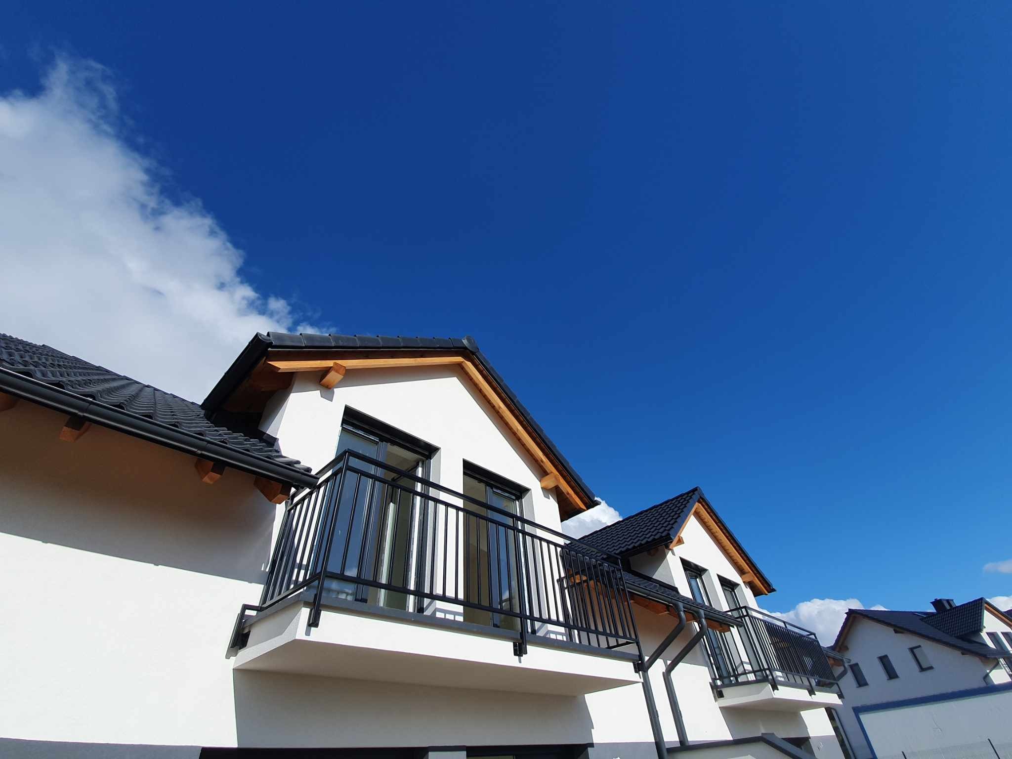 Nowoczesna balustrada balkonowa z czarnych, pionowych prętów na tle białej elewacji budynku i błękitnego nieba z chmurami.