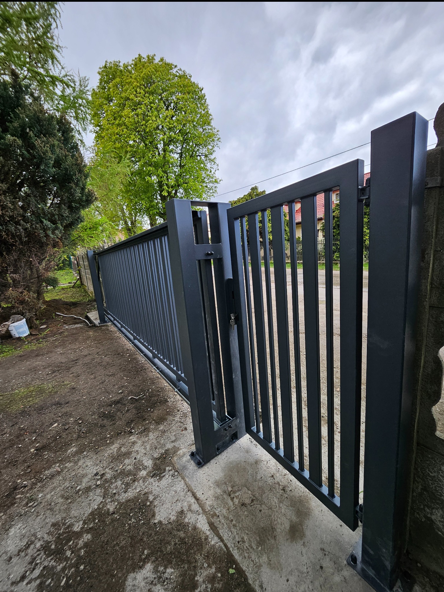 Nowoczesna, częściowo otwarta, przesuwna brama wjazdowa w kolorze grafitowym, z pionowymi szczeblami, zamontowana na betonowym podłożu, z widocznym mechanizmem otwierania i zielenią w tle.