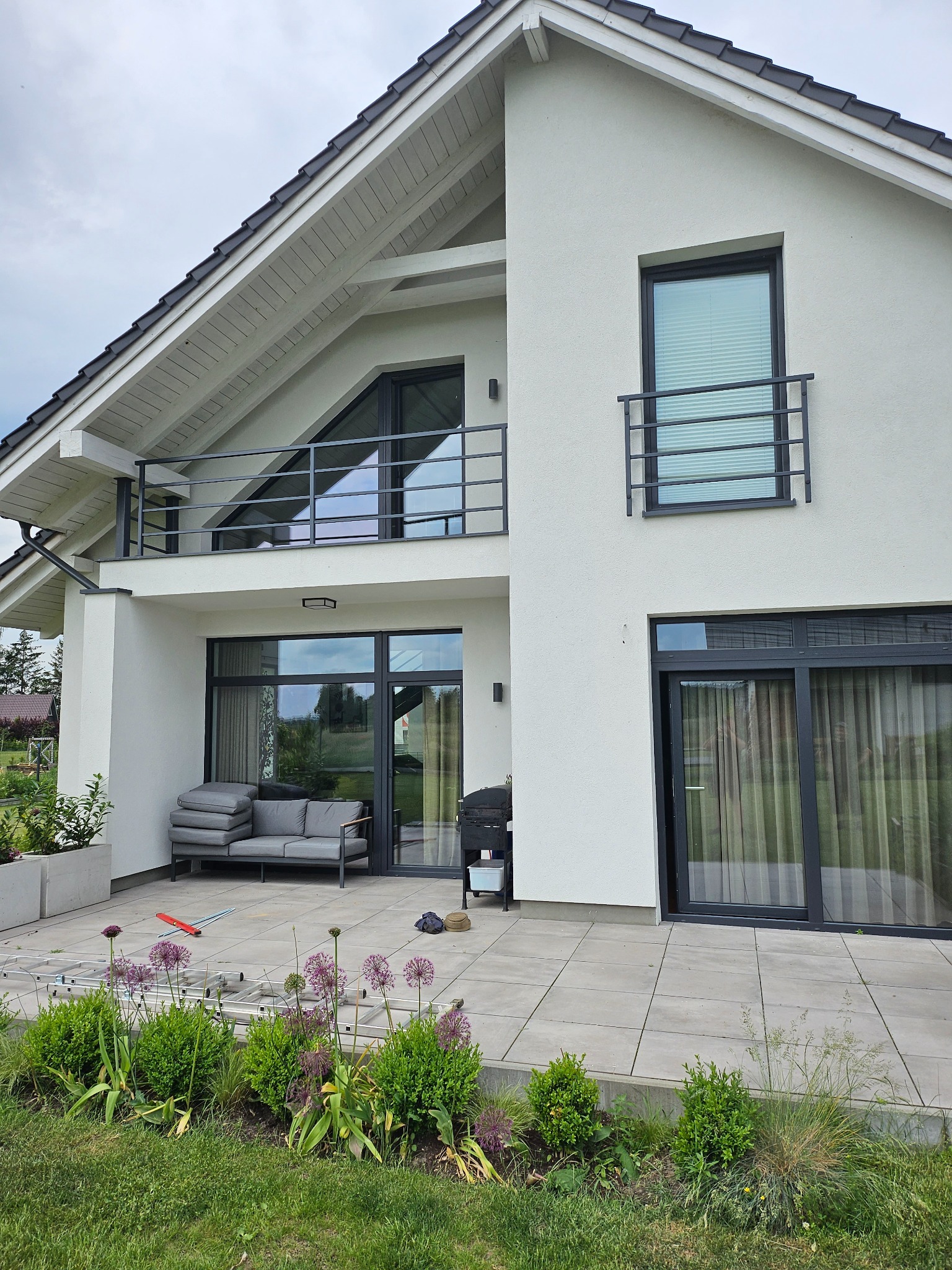 Nowoczesny dom z białym tynkiem, balkonem z czarną balustradą i tarasem wyłożonym płytami, widok z ogrodu na fragment elewacji z czarnymi ramami okiennymi i rozłożoną drabiną na tarasie.