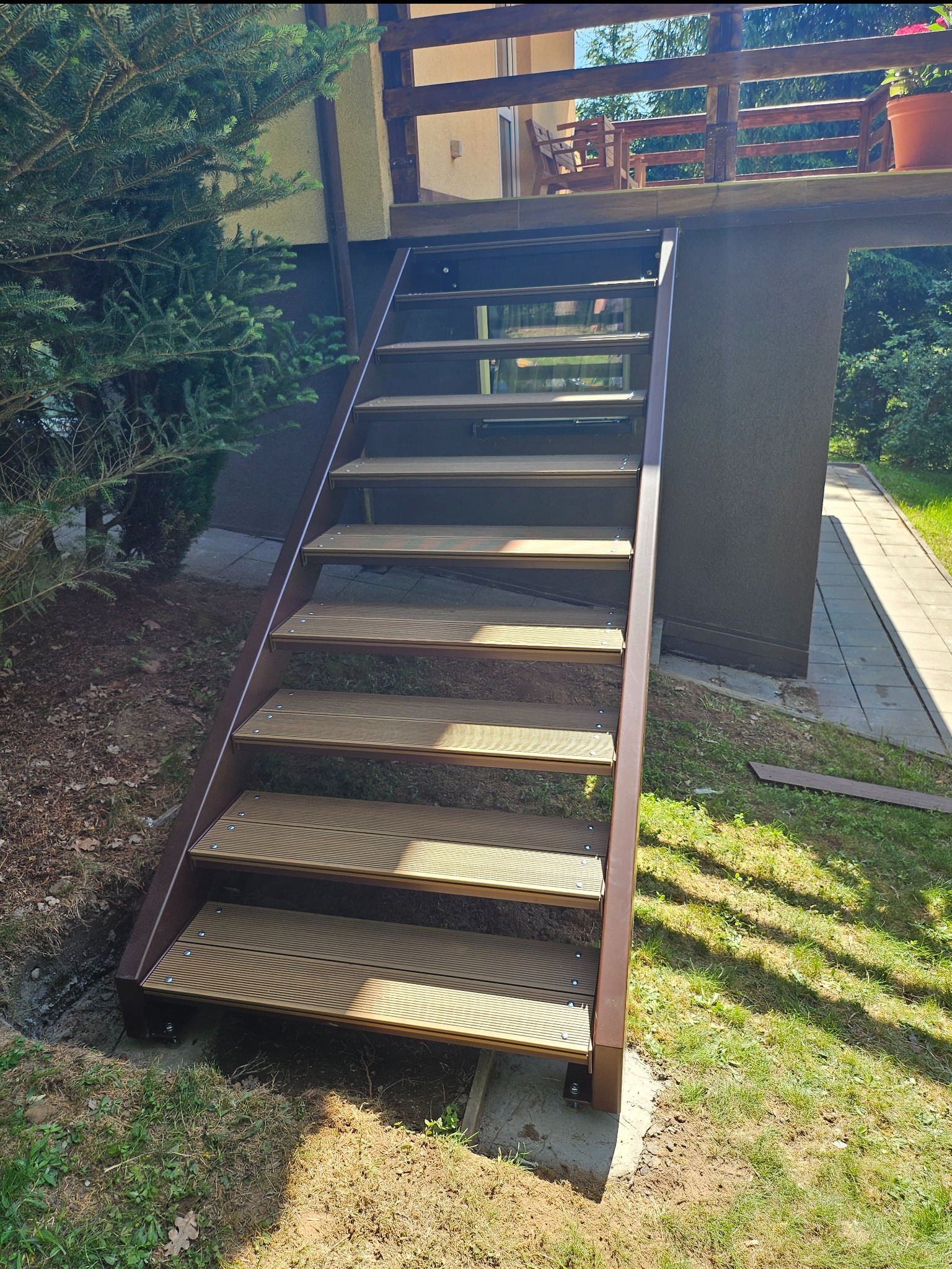 Zewnętrzne schody z brązową metalową ramą i deskami kompozytowymi, prowadzące na balkon domu otoczonego zielenią.