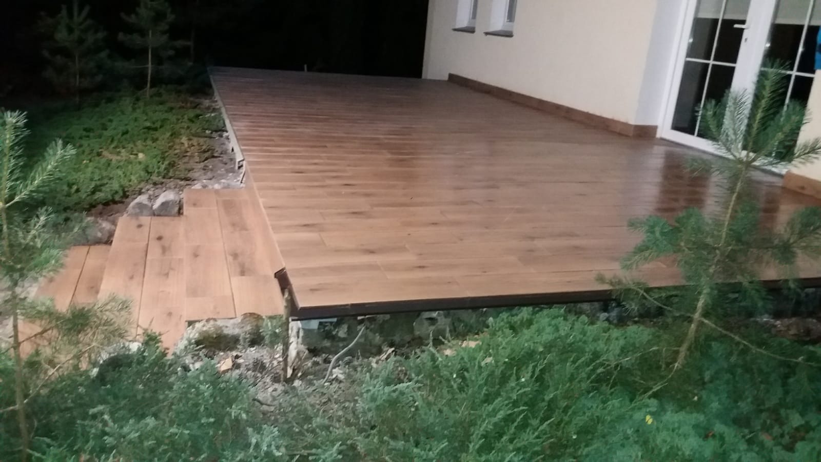 Wykończony taras z płytek imitujących drewno, z niskimi schodami prowadzącymi do ogrodu, widok z poziomu gruntu, oświetlenie wieczorne.