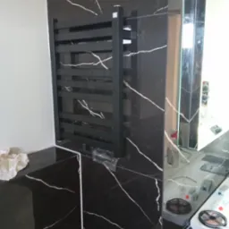 Czarny grzejnik drabinkowy na ścianie z czarnego marmuru z białymi żyłami, widoczne elementy instalacji i fragment lustra.