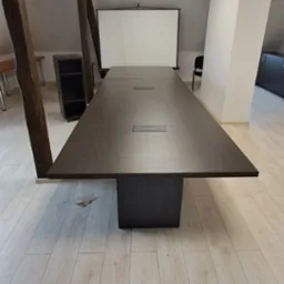 Długa, ciemnobrązowa drewniana stół konferencyjny z wbudowanymi gniazdami, umieszczona w jasnym pomieszczeniu z drewnianymi podłogami i projektorem ekranowym w tle, wsparta na solidnej, ciemnej...