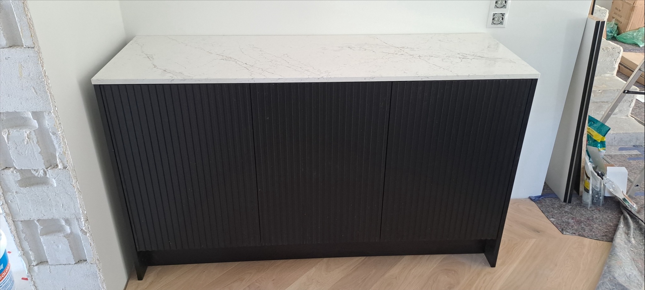 Czarna szafka z ryflowanymi drzwiczkami i marmurowym blatem w trakcie montażu, widoczne materiały budowlane i surowa ściana z cegieł.