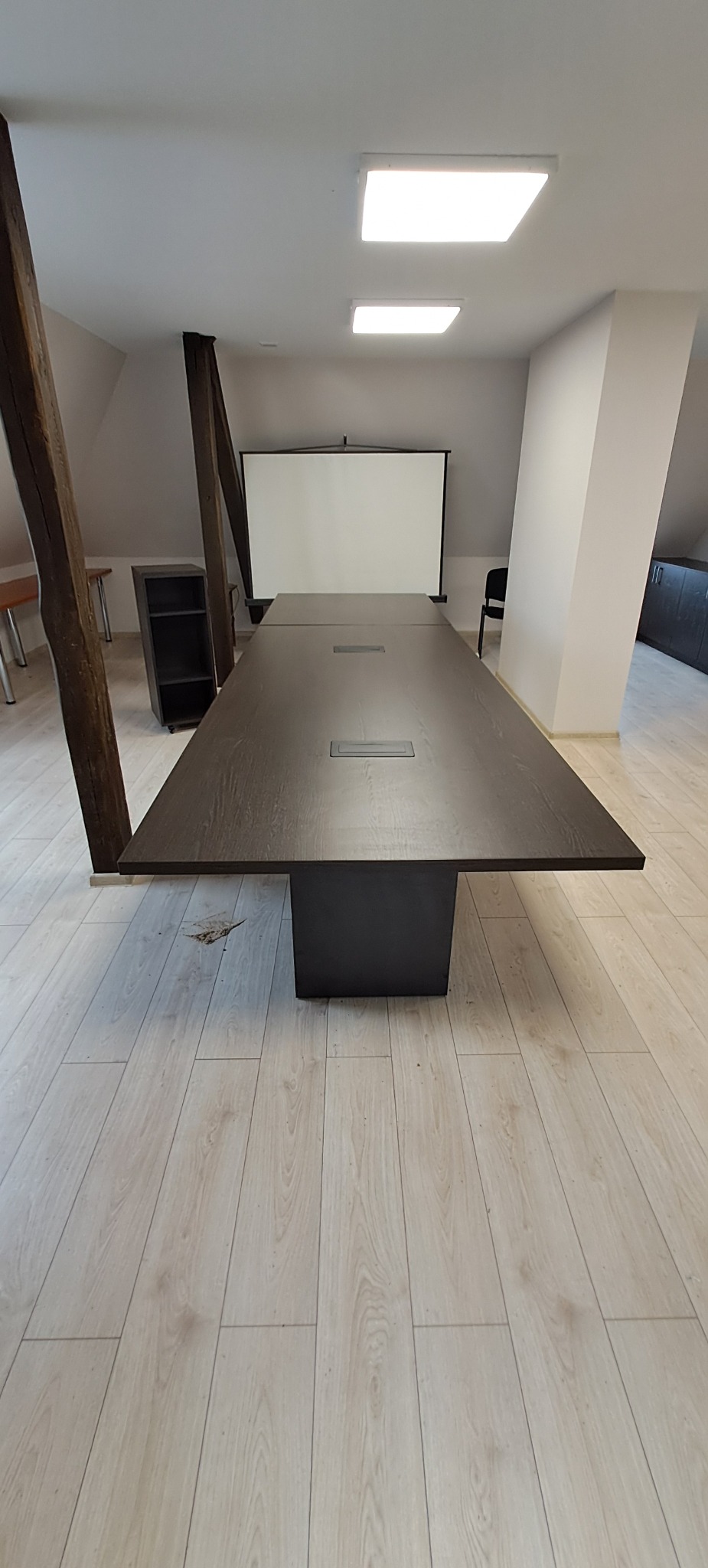 Długa, ciemnobrązowa drewniana stół konferencyjny z wbudowanymi gniazdami, umieszczona w jasnym pomieszczeniu z drewnianymi podłogami i projektorem ekranowym w tle, wsparta na solidnej, ciemnej...