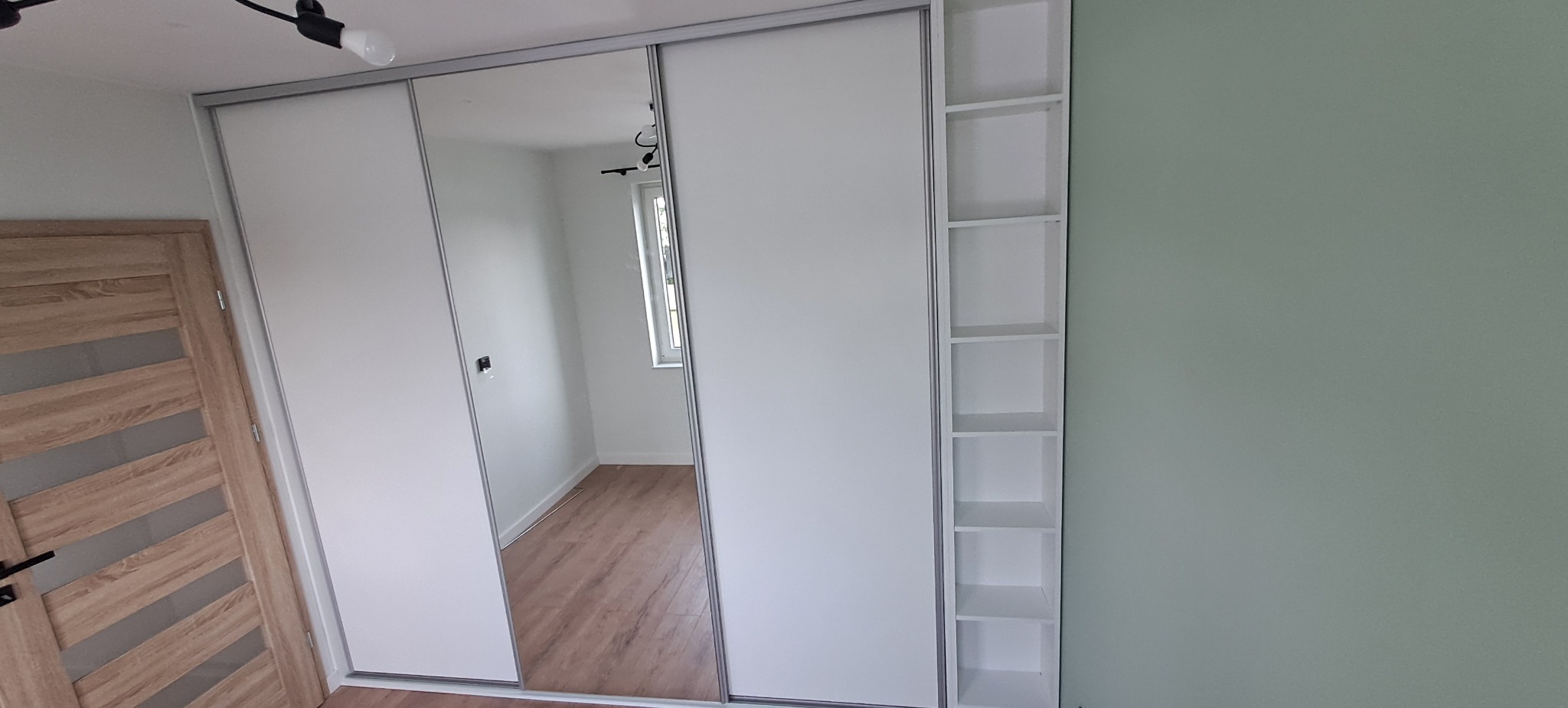 Biała szafa przesuwna z lustrem i półkami bocznymi w jasnym pokoju, widoczna drewniana okleina drzwi po lewej stronie.