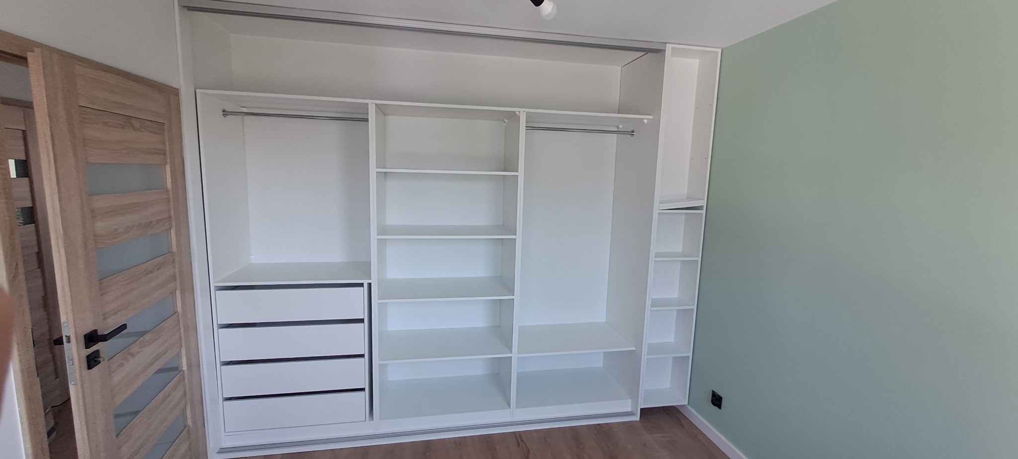 Biała szafa wnękowa z przesuwnymi drzwiami, z szufladami i półkami, zamontowana przy ścianie w kolorze miętowym, widoczny fragment drewnianych drzwi z przeszkleniami.