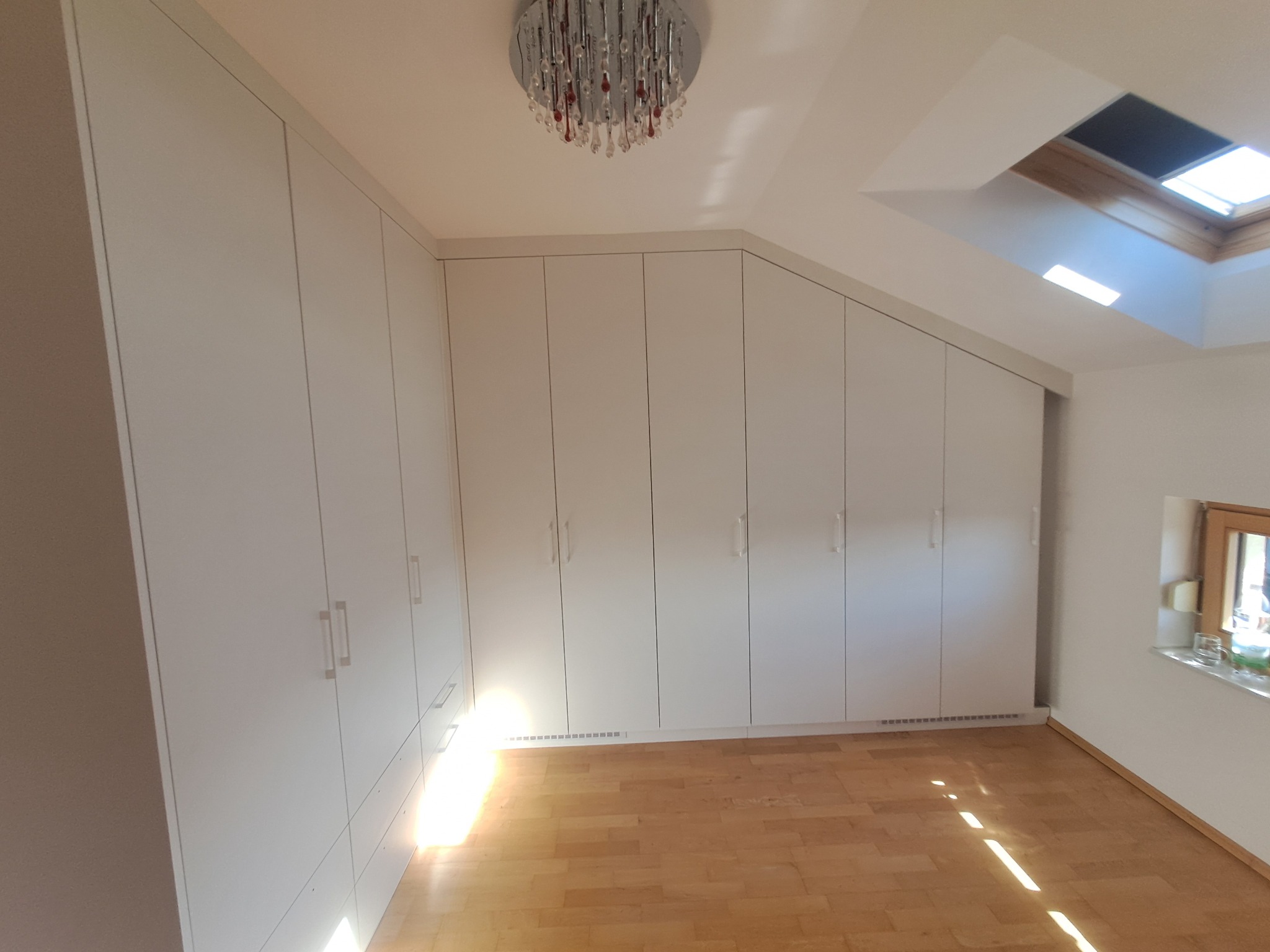 Białe szafy wnękowe pod skosem dachu, z oświetleniem punktowym i parkietem na podłodze.