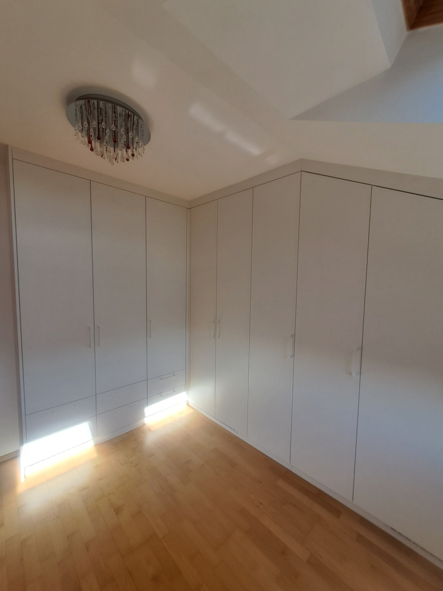 Białe szafy wnękowe pod skosem dachu, z drewnianą podłogą i ozdobnym plafonem sufitowym.