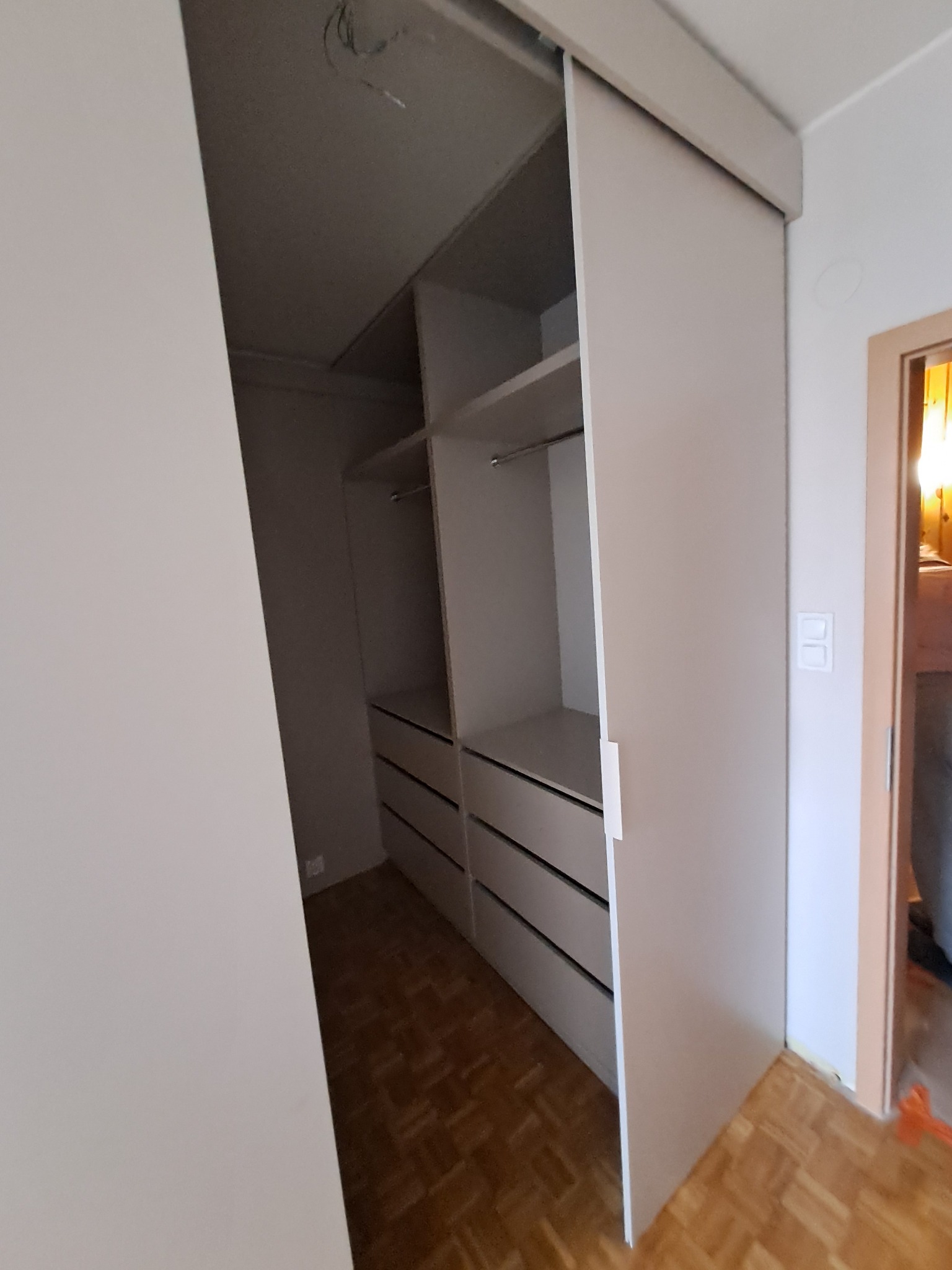 Wnętrze szafy wnękowej z przesuwnymi drzwiami, widoczne półki i szuflady, jasne kolory, parkiet na podłodze.