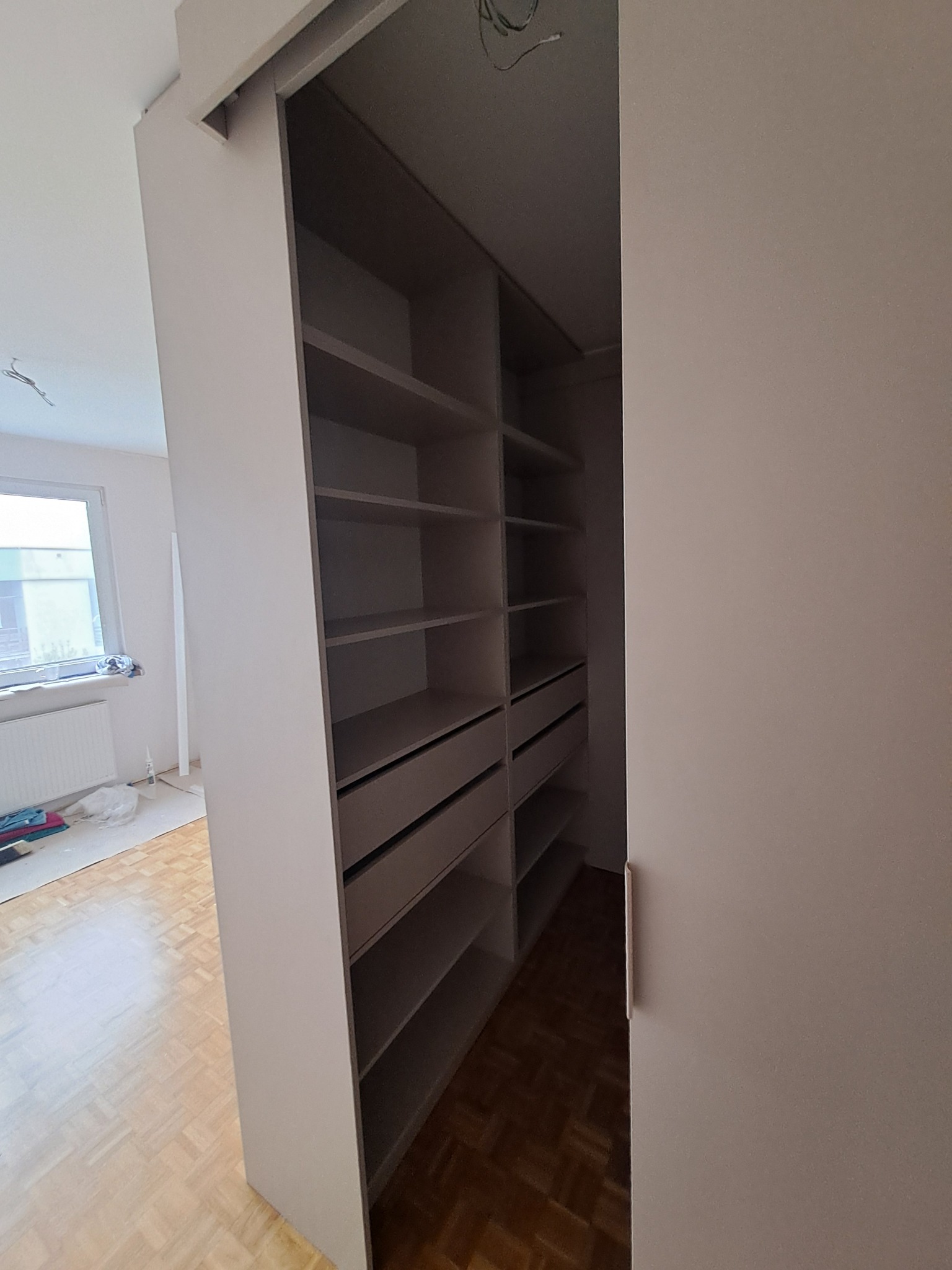 Wnętrze szafy wnękowej z półkami i szufladami, widoczne przesuwne drzwi i fragment pokoju z parkietem i oknem.